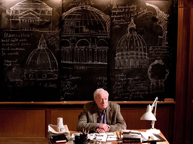 Michael Caine sitzt vor einer Tafel mit Zeichnungen des Petersdoms.