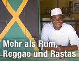 Ein Jamaikaner steht vor offenem Fenster mit aufgemalter Jamaika-Flagge auf den Fensterläden