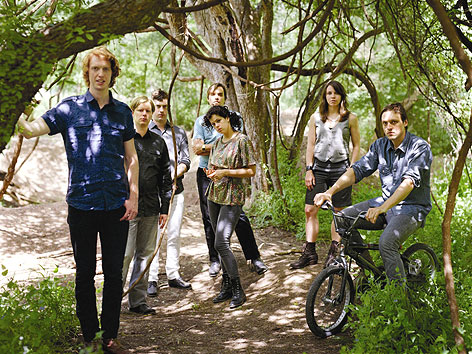 © Eric Kayne Mitglieder der Band "Arcade Fire" stehen mit einem Fahrrad im Wald.
