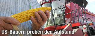 Amerikanischer Farmer mit einem Maiskolben in der Hand und einem M&auml;hdrescher im Hintergrund