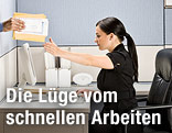 Eine Frau arbeitet im Büro am Computer und bekommt gleichzeitig, ohne hin zu sehen, einen Akt in die Hand gedrückt