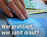 Eine Hand liegt auf einem Sparbuch (Euroscheine im Hintergrund).