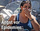 Frau vor einem Müllberg hält sich die Hand vor ihrem Mund