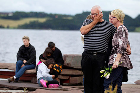 Ein Mann und eine Frau mit Blumen in der Hand weinen am Ufer vis a vis der Insel Utoya