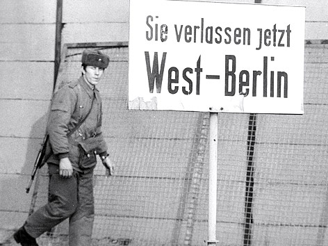 © AP/Edwin Reichert Schwarzweißaufnahme eines ostdeutschen Grenzpolizisten in Berlin neben einem Schild mit der Aufschrift "Sie verlassen jetzt West-Berlin"