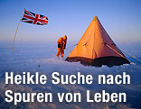 Eine Person steht mit Kapuze und Anorak vor einem Zelt in der Antarktis und neben der britischen Flagge.