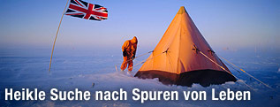 Eine Person steht mit Kapuze und Anorak vor einem Zelt in der Antarktis und neben der britischen Flagge.