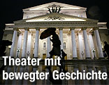 Eine Person mit Regenschirm geht nachts am beleuchteten Bolschoi-Theater in Moskau vorbei.