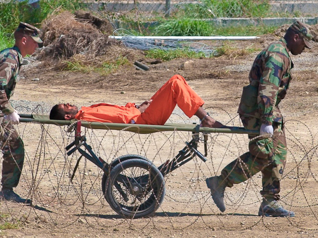 © AP/Lynne Sladky Ein Gefangener in einem orangen Anzug wird von zwei Soldaten auf einem Rollwagen zu einer Verhörung geführt