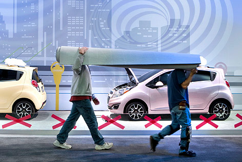 Arbeiter tragen einen Teppich an zwei ausgestellten Chevrolet-Kleinwagen vorbei