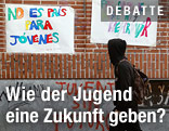 Junger Mann mit Kapuzenpullover geht an einem Schild mit der spanischen Aufschrift "No es pais para jovenes" ("Jugend ohne Zukunft") vorbei