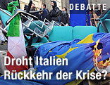 Eine hängende italienische Flagge neben einer brennenden EU-Flagge vor vermummten Demonstranten
