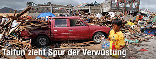 Ein Bub geht mit einer Wasserflasche an zerstörten Häusern und einem beschädigten Auto vorbei