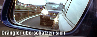 Im Rückspiegel eines Auto ist knapp dahinter ein anderes Auto mit Licht zu sehen