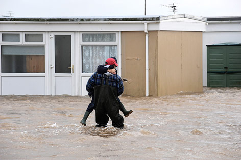 © Reuters/Rebecca Naden Eine Person wird in Kidwelly in Wales von einem Helfer über eine überflutete Straße getragen