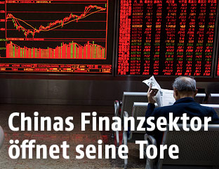 Mann sitzt mit einer Zeitung vor den Aktienkursen der Pekinger Börse