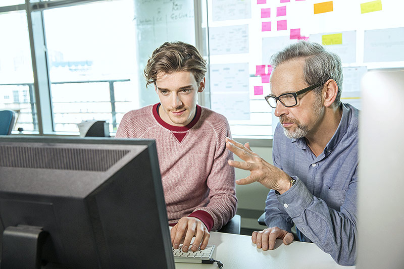 © Getty Images/Igor Emmerich Ein Mann mit grauem Bart schult einen jungen männlichen Praktikanten vor einem PC-Monitor ein